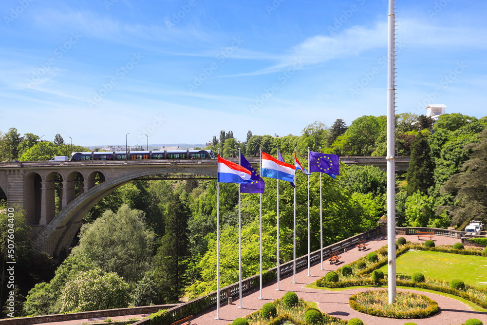 View of the Adolphe Bridge in Luxembourg City. Flags of Luxembourg and the European Union fly in the foreground - obrazy, fototapety, plakaty 