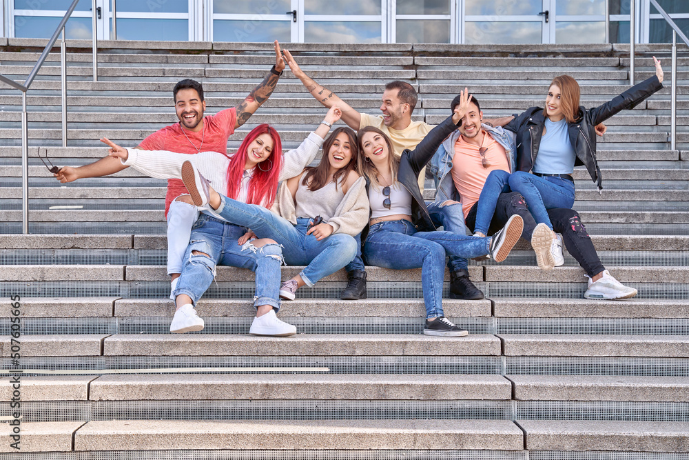 Group of happy young friends having fun and smiling at camera while sitting together on stairs outdoors. Friendship and positivity concept.