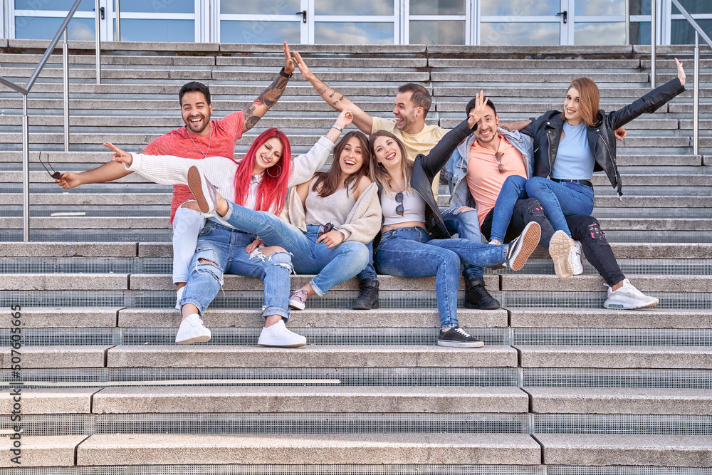 Obraz premium Group of happy young friends having fun and smiling at camera while sitting together on stairs outdoors. Friendship and positivity concept.