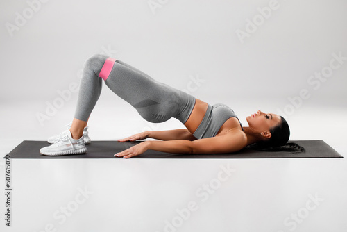 Athletic girl doing glute bridge exercise with resistance band on gray background. Fitness woman working out.