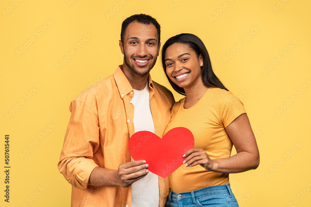 Romantic young black couple with red paper heart in hands posing over yellow studio background Romantic young black couple with red paper heart in hands posing over yellow studio background