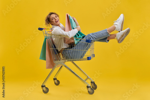 Delighted woman hugging bright paper bags, while sitting in shopping trolley. Side view of happy girl cuddling purchases, looking at camera, isolated on yellow studio background. Concept of shopping.