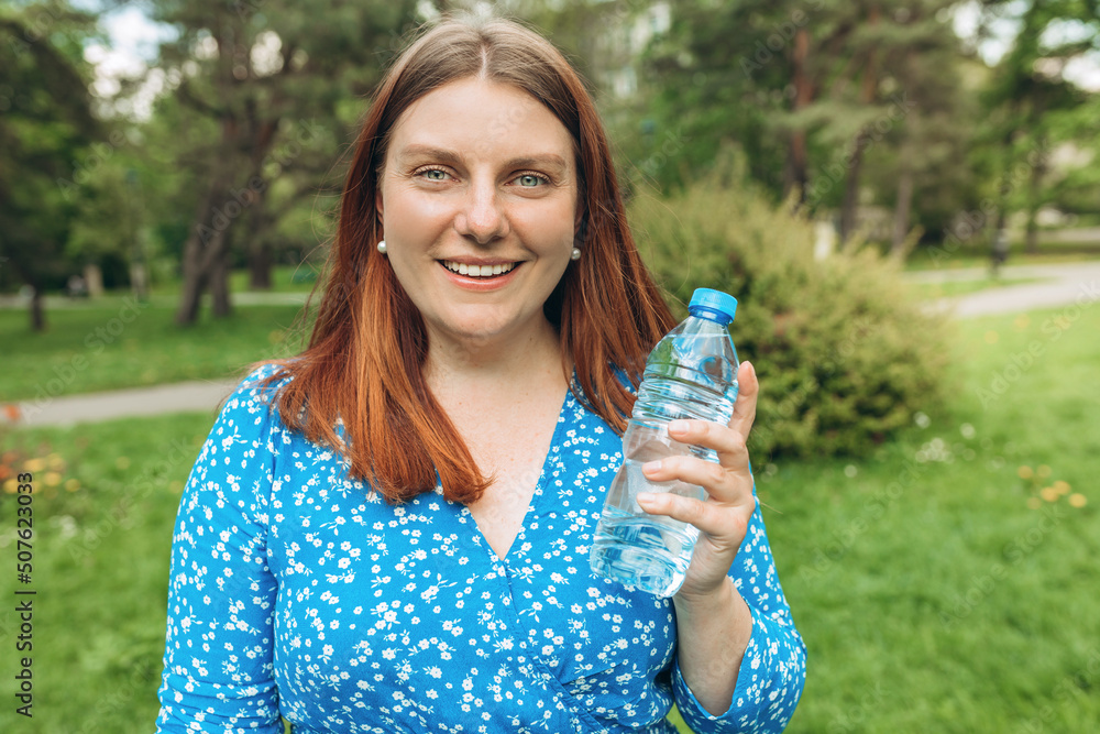 Beautiful happy young woman holding water bottle against green nature background. Optimistic redhead girl in blue dress walking in city summer park, Healthy concept. Beautiful happy young woman holding water bottle against green nature background. Optimistic redhead girl in blue dress walking in city summer park, Healthy concept.