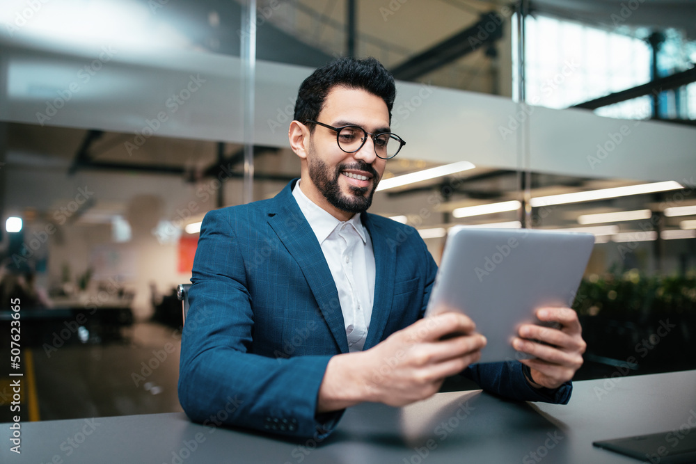 Smiling busy handsome millennial middle eastern businessman with beard in glasses, suit works with tablet Smiling busy handsome millennial middle eastern businessman with beard in glasses, suit works with tablet