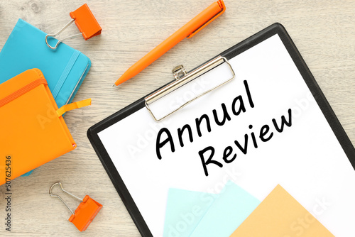 ANNUAL REVIEW. text on clipboard folder, orange sticker and blue and orange notepad.