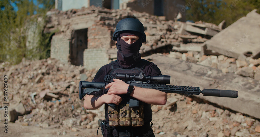 Fototapeta premium A soldier with a machine gun stands against the background of a destroyed house, looks around and raises his weapon.