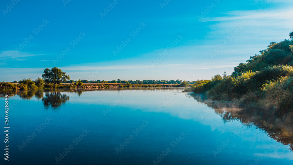 Fototapeta premium Aerial view of a beautiful summer landscape over river while dawn. Top view over river with a smooth water surface reflecting blue sky.
