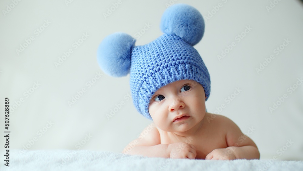 Sweet newborn baby in a blue hat with pompoms looks into the camera. A calm two-month-old infant is lying on a blanket. A beautiful, cute little child with blue eyes looks into the camera. . Sweet newborn baby in a blue hat with pompoms looks into the camera. A calm two-month-old infant is lying on a blanket. A beautiful, cute little child with blue eyes looks into the camera. .