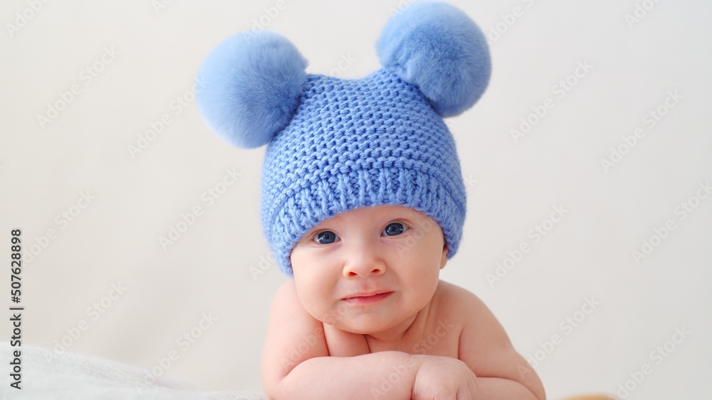 Sweet newborn baby in a blue hat with pompoms looks into the camera. A calm two-month-old infant is lying on a blanket. A beautiful, cute little child with blue eyes looks into the camera. . Sweet newborn baby in a blue hat with pompoms looks into the camera. A calm two-month-old infant is lying on a blanket. A beautiful, cute little child with blue eyes looks into the camera. .