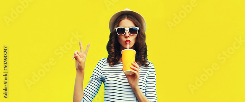 Portrait of stylish young woman drinking fresh juice wearing summer straw hat, sunglasses and striped t-shirt on yellow background