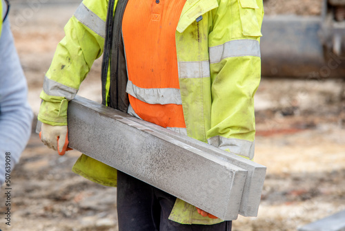 Close up of groundworker in orange and yellow hi-viz  carrying heavy concrete kerbs on construction site during new road construction photo