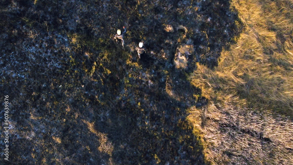 Two firefighters walk on black scorched earth after fire and burning dry grass in field. Two people in uniforms of firefighters walk on black earth and ash after extinguishing fire. Aerial drone view Two firefighters walk on black scorched earth after fire and burning dry grass in field. Two people in uniforms of firefighters walk on black earth and ash after extinguishing fire. Aerial drone view