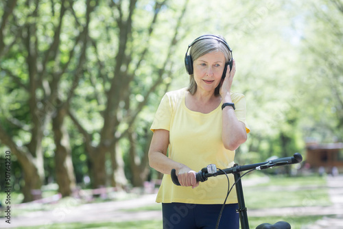 Active lifestyle. Senior beautiful woman walks in the park, rides a yellow bike, listens to music on headphones. Standing with a bicycle in his hands, smiling, having a good time