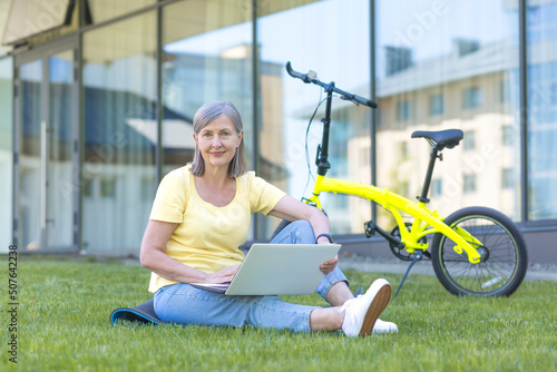 Teaching. Portrait of senior beautiful woman sitting on campus on grass with laptop and bicycle. He studies and works. He looks at the camera, smiles