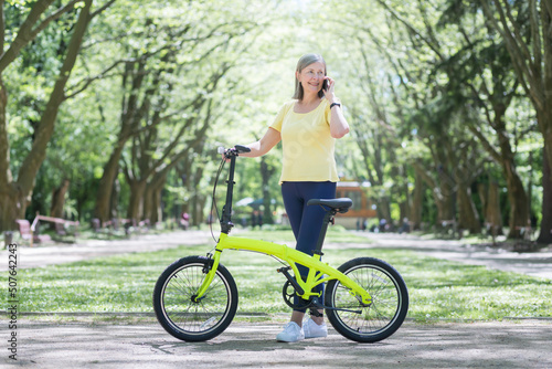 Active lifestyle. Senior beautiful woman walks in the park, rides a yellow bike, talks on the phone. Standing with a bicycle in his hands, smiling, having a good time © Liubomir