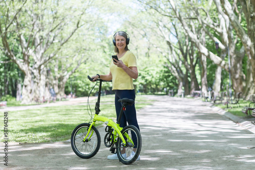 Senior beautiful woman walks in the park, rides a bike, listens to music on headphones, holds a phone. He stands with a bicycle in his hands, looks at the camera, smiles.