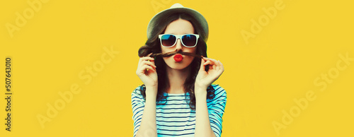 Portrait of funny young woman showing mustache her hair blowing lips with red lipstick sending air kiss on colorful yellow background, blank copy space for advertising text