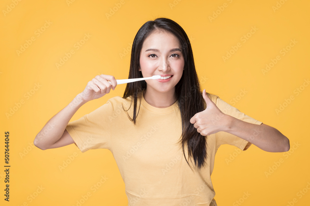 Portrait of Happy Asian female brushing her Teeth. Young Asian Woman brushing her teeth with clean white teeth. Happy Asian Woman wearing Yellow T-shirt standing over yellow background. Portrait of Happy Asian female brushing her Teeth. Young Asian Woman brushing her teeth with clean white teeth. Happy Asian Woman wearing Yellow T-shirt standing over yellow background.