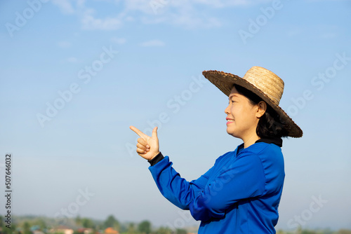 Side view of Asian woman famer, wears hat, blue shirt, raise finger up to point on sky background. Copy space for adding text or advertisement. Concept : Agriculture occupation. Happy farmer.