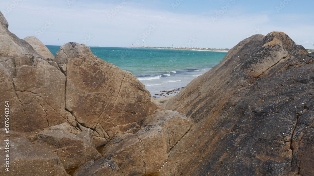Tas de gros rochers sur la belle plage de Plouédern en Bretagne, avec un mer bleu turquoise, de la végétation maritime verte et des flaques d'eau, et des vagues