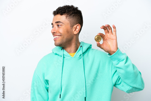 Young Brazilian man holding a Bitcoin isolated on white background looking side