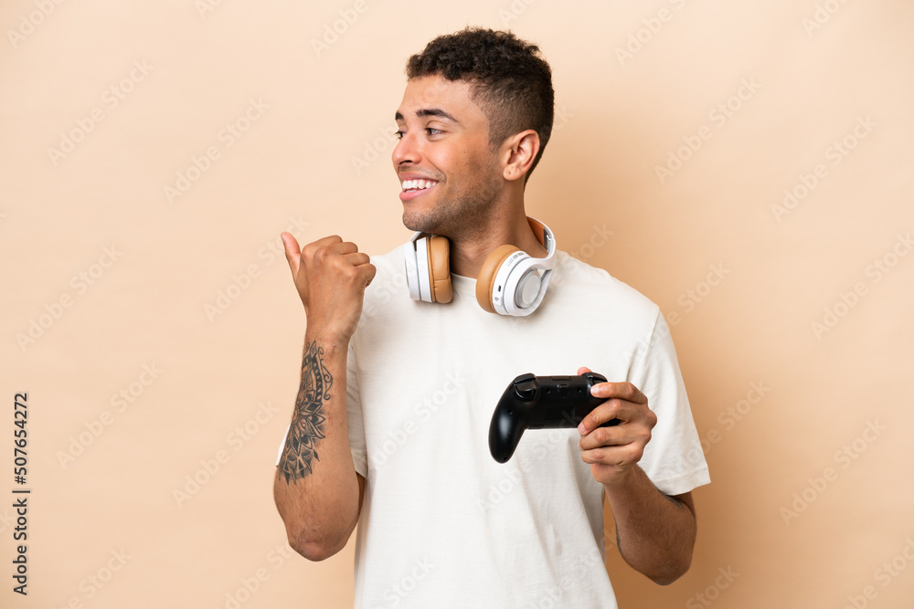 Young Brazilian man playing with a video game controller isolated on beige background pointing to the side to present a product Young Brazilian man playing with a video game controller isolated on beige background pointing to the side to present a product