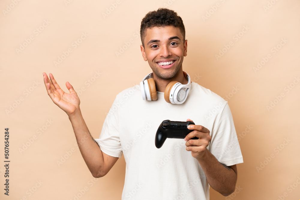 Young Brazilian man playing with a video game controller isolated on beige background extending hands to the side for inviting to come Young Brazilian man playing with a video game controller isolated on beige background extending hands to the side for inviting to come