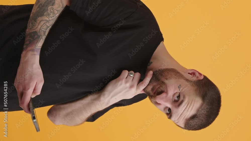 Vertical video attracted tattooed man swipes on his phone, looks at the camera and smiles yellow background isolated studio shot . High quality 4k footage Vertical video attracted tattooed man swipes on his phone, looks at the camera and smiles yellow background isolated studio shot . High quality 4k footage