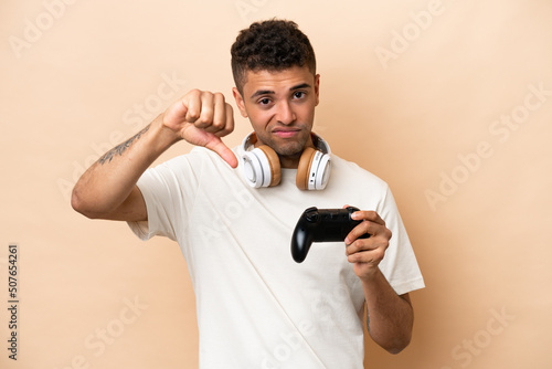 Young Brazilian man playing with a video game controller isolated on beige background showing thumb down with negative expression