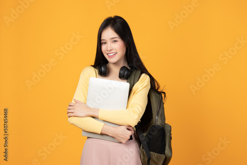 Portrait of happy casual Asian girl student with backpack, headphones and laptop isolated on yellow background. Back to school and learning concept.