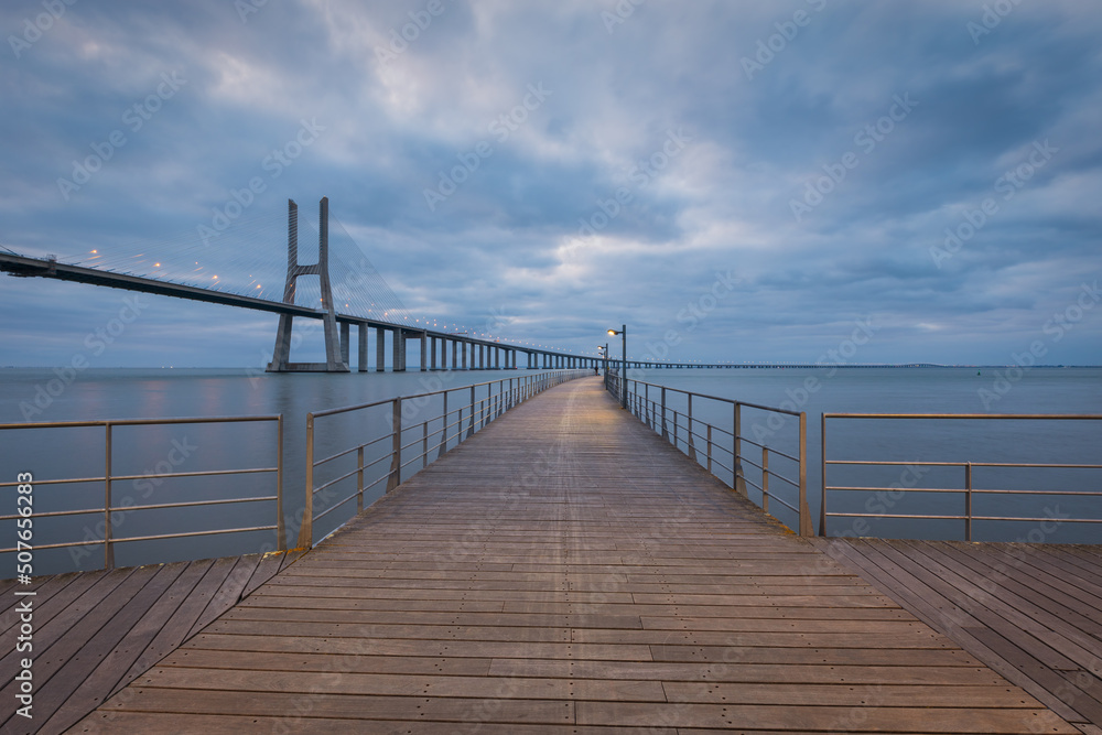 Vasco da Gama bridge and pier over tagus river in Lisbon, Portugal, at dawn, with a cloudy sky.