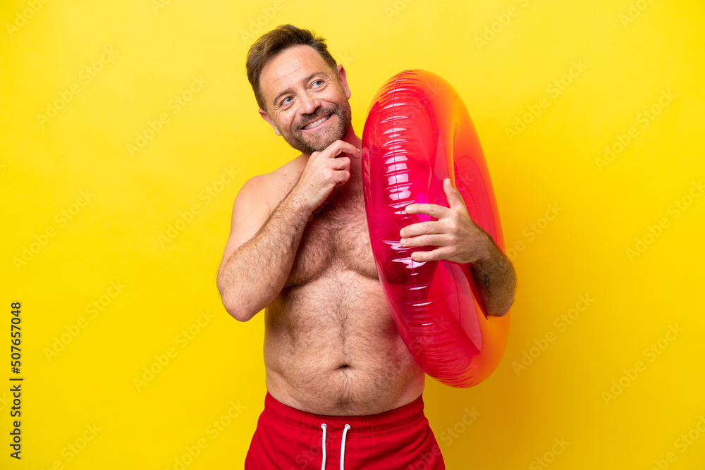 Middle age caucasian man holding inflatable donut isolated on yellow background looking up while smiling