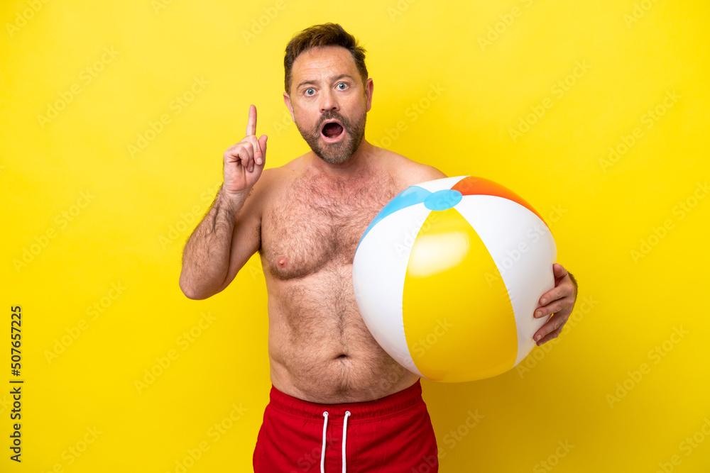 Middle age caucasian man holding beach ball isolated on yellow background thinking an idea pointing the finger up