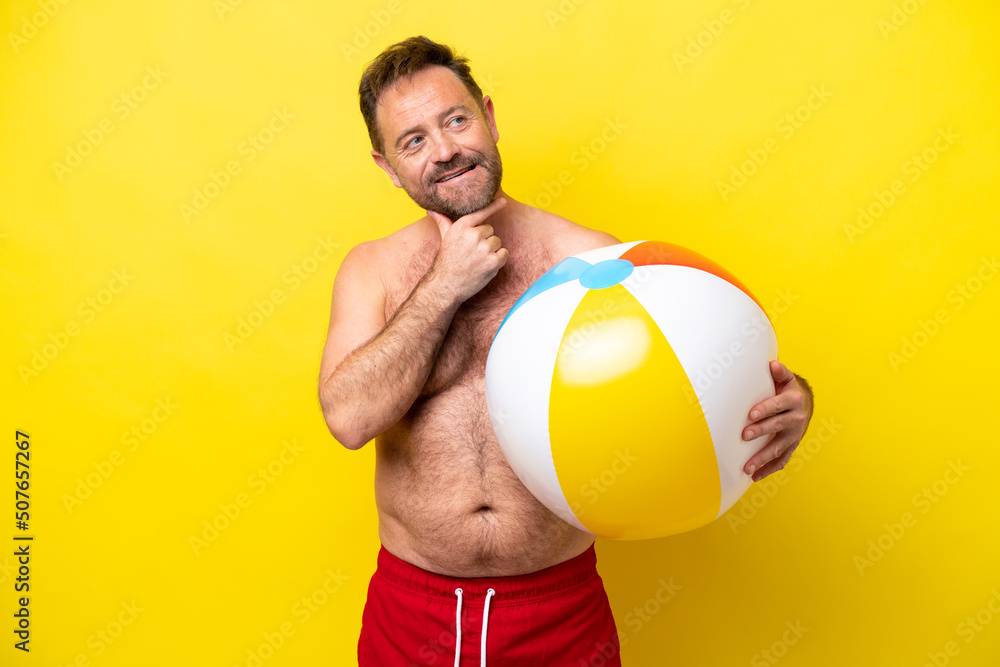Middle age caucasian man holding beach ball isolated on yellow background looking up while smiling