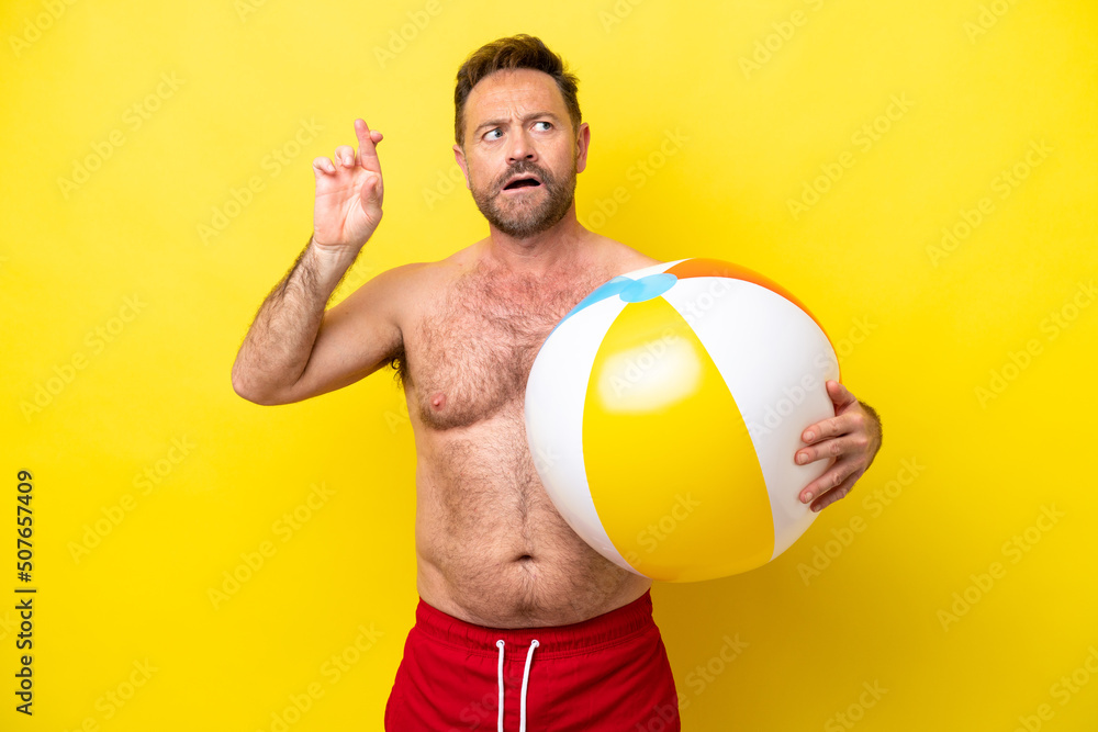 Middle age caucasian man holding beach ball isolated on yellow background with fingers crossing and wishing the best