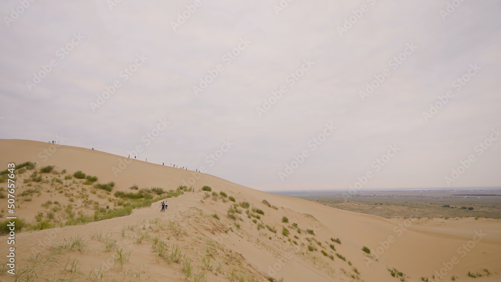 People walk along sandy ridge. Action. Tourists walk on sand hills in desert. Walking tour in desert on background of endless horizon in cloudy weather