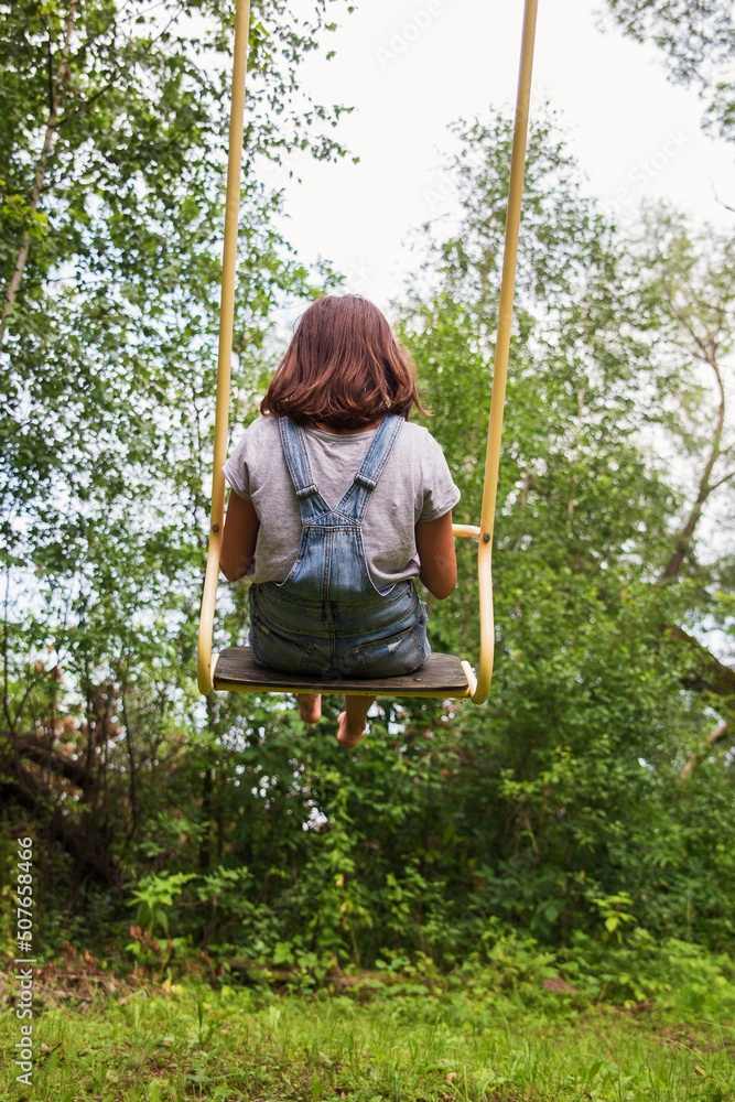 a school-age girl in blue denim shorts and a gray t-shirt rides on a swing in the garden of summer, back view
