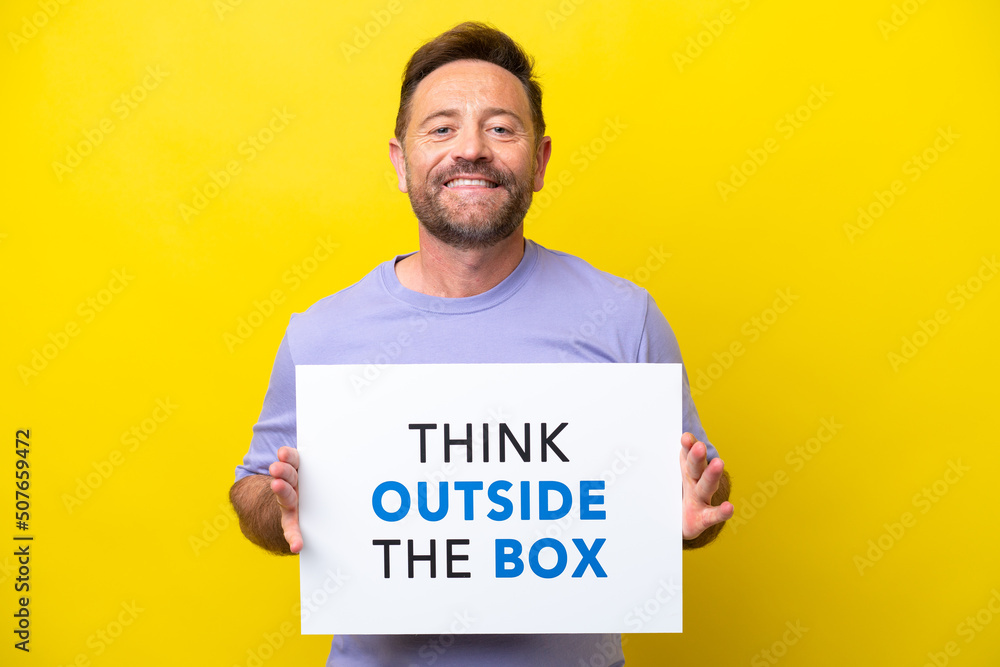 Middle age caucasian man isolated on yellow background holding a placard with text Think Outside The Box Middle age caucasian man isolated on yellow background holding a placard with text Think Outside The Box