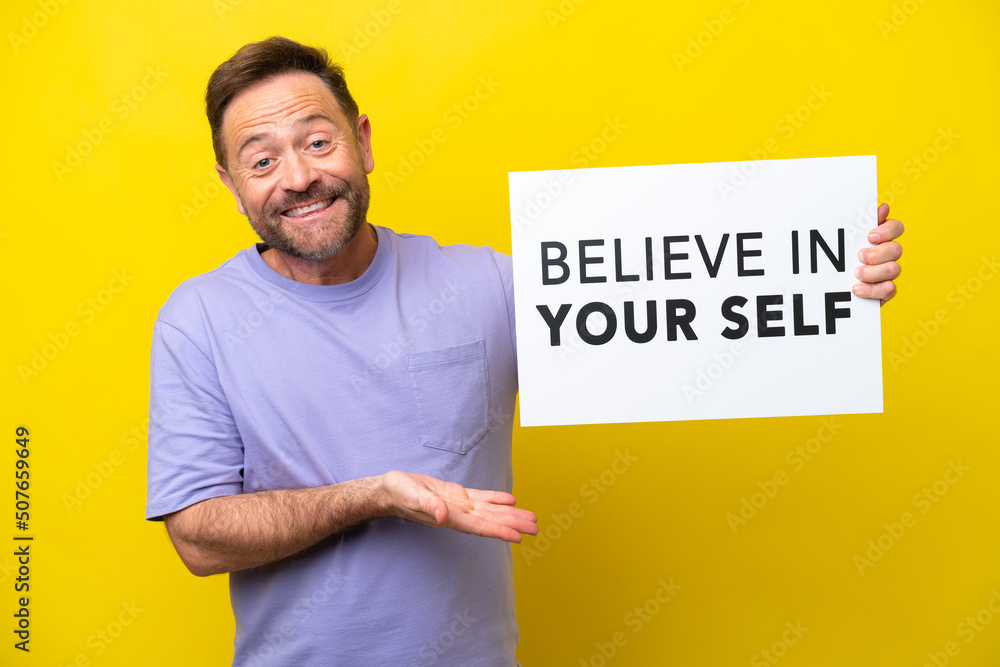 Middle age caucasian man isolated on yellow background holding a placard with text Believe In Your Self and pointing it Middle age caucasian man isolated on yellow background holding a placard with text Believe In Your Self and pointing it