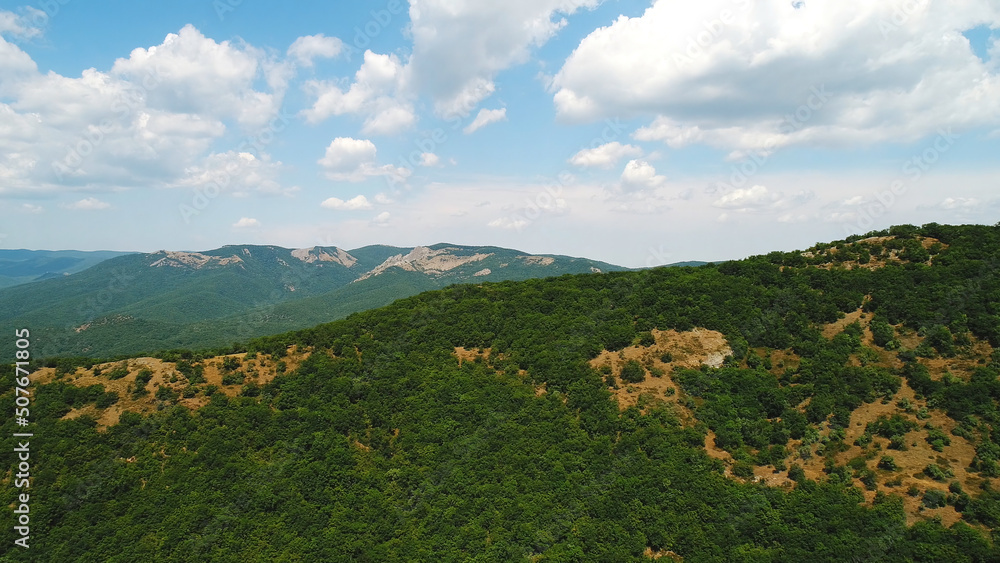 Aerial view of the grey mountain ranges and mountain slopes covered by green trees and shrubs against the blue cloudy sky in summer day. Shot. Beautiful mountain scenery