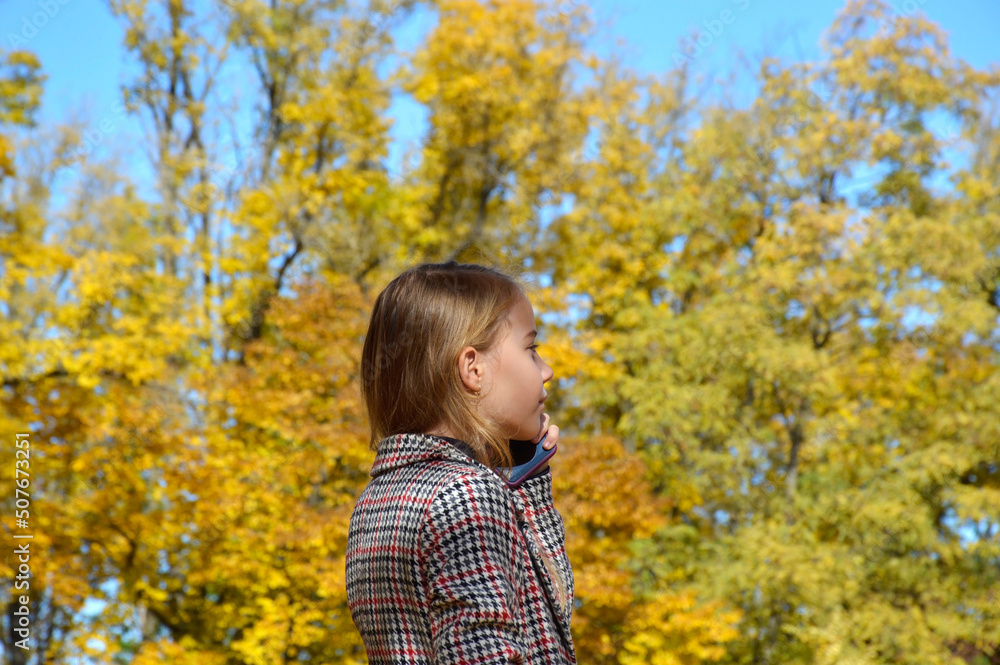 beautiful girl in a plaid coat talking on the phone in the autumn park. the child enjoys the warm rays and the beauty of autumn nature beautiful girl in a plaid coat talking on the phone in the autumn park. the child enjoys the warm rays and the beauty of autumn nature
