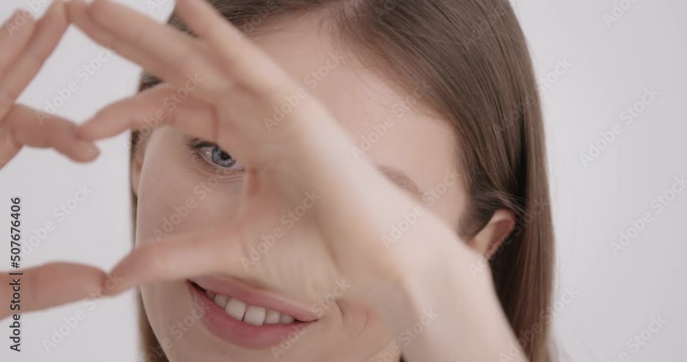 Healthy eyes and vision. Portrait of young woman making heart shape gesture while smiling and looking at camera in studio. Caucasian female model with healthy fresh skin over grey background. Healthy eyes and vision. Portrait of young woman making heart shape gesture while smiling and looking at camera in studio. Caucasian female model with healthy fresh skin over grey background.
