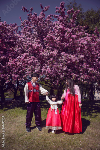 Korean family in national costumes in nature stands next to a cherry blossoming tree.