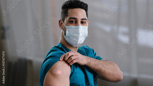 Inspired arabian hispanic man young male practitioner nurse intern in medical face mask showing injection mark adhesive bandage shoulder make vaccine from covid-19 virus pandemic coronavirus infection