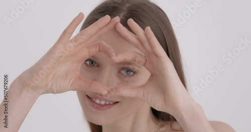 Healthy eyes and vision. Gorgeous young woman with natural beauty showing heart shape with hands over grey studio background. Skin care, love and feelings concept.