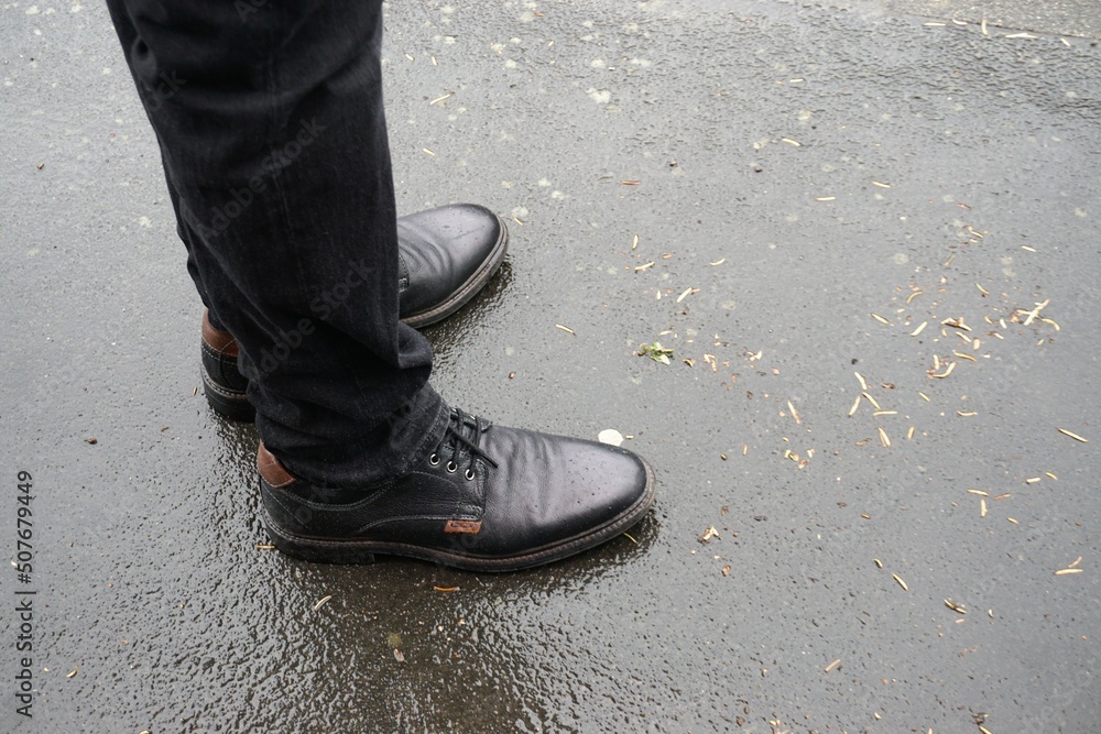 Schwarzes Paar Lederschuhe mit schwarzen Hosenbeinen von Mann auf nasser Straße bei Regen im Frühling 