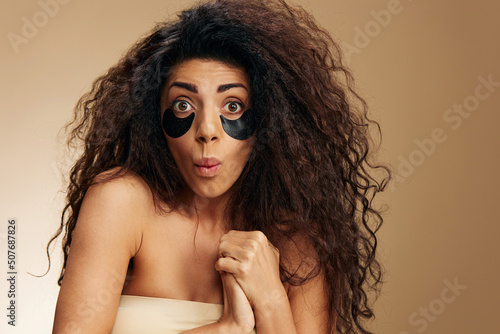 Fotografiet Excited pretty Latin female with black hydrogel patches under eye say Wow folded hands posing isolated over pastel beige background