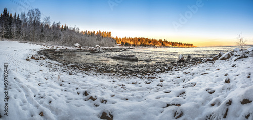 A panoramic sunset horizon view of winter landscape seashore with snow covered pine trees