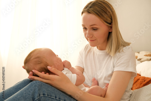 mom holds a newborn baby in her arms. in the background the sun shines from the window.