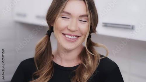 Close-up indoor portrait of a charming and a bit shy dark blond woman. Pretty girl smiling when looking into the camera and touching her pigtails